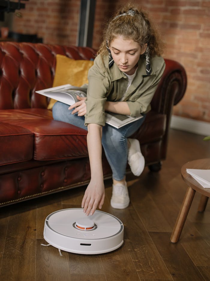 A young woman relaxes on the couch with a book while a robot vacuum cleans the floor.